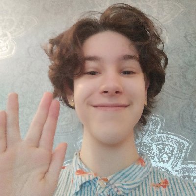 A white non binary person doing the vulcan salute. They are wearing a blue and white shirt that's patterned with fish, and their hair is brown and wavy.